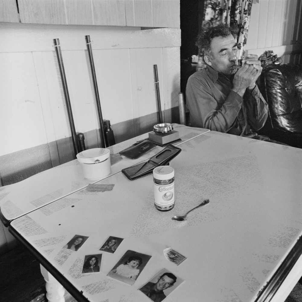 A man sits at a table, smoking. The table holds a tin of coffee, an ashtray, a spoon, and several photographs. A patterned sofa is visible in the background. The scene is in black and white.