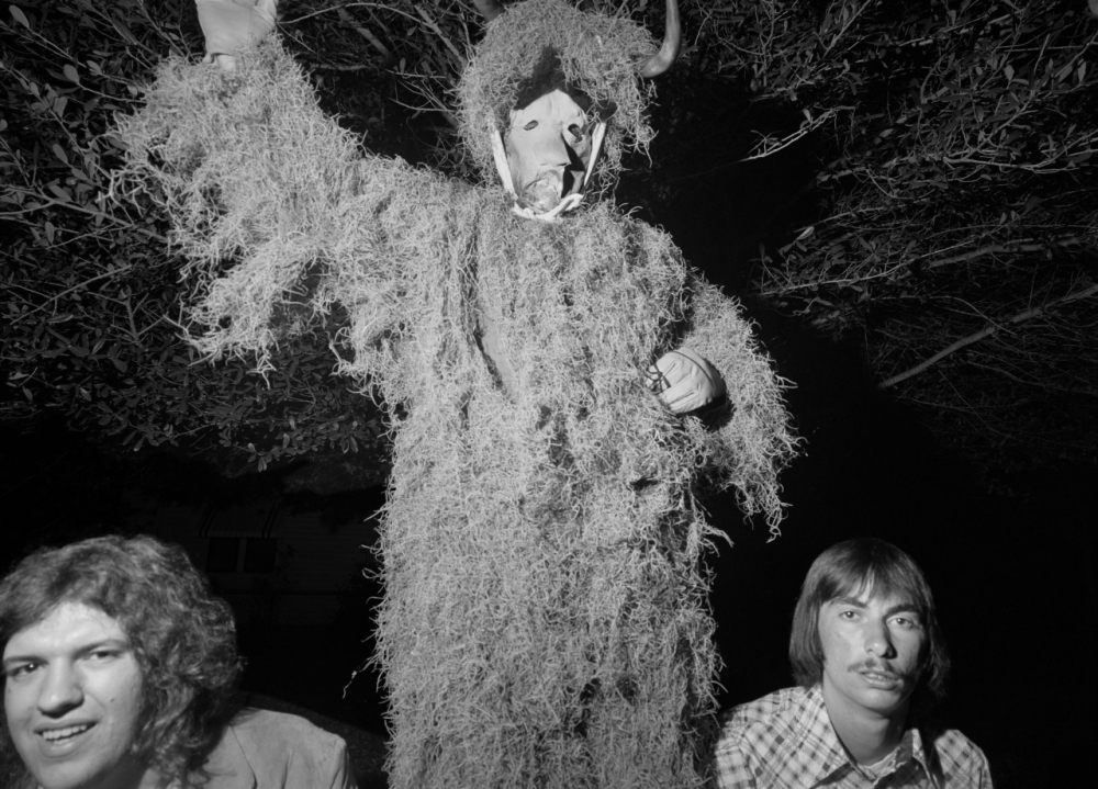 A person in a costume covered in moss or foliage, wearing a horned mask, stands outdoors at night. Two men stand nearby, visible from the shoulders up. One has curly hair; the other has straight hair and a mustache. Trees form the backdrop.