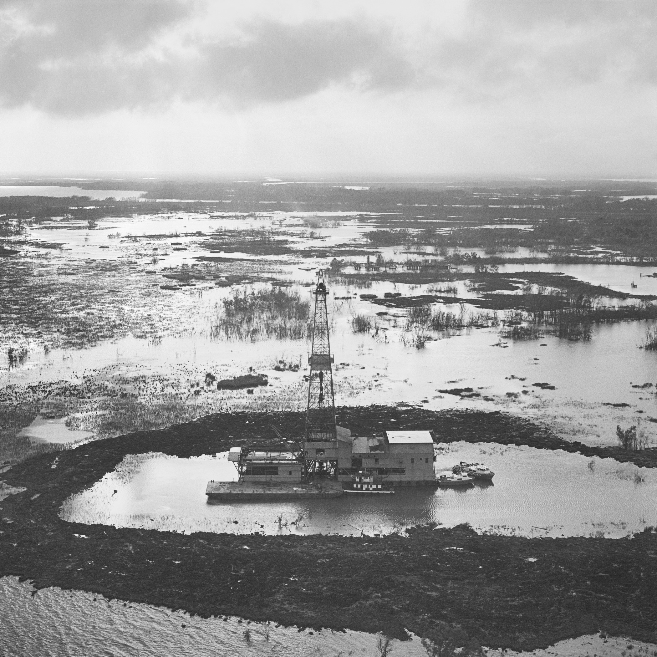 An aerial view of an offshore oil drilling rig surrounded by waterlogged, marshy terrain. The landscape appears flooded, with sparse trees and vegetation visible amidst the waterlogged area under a cloudy sky.