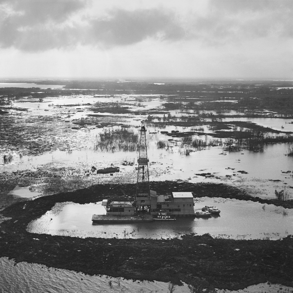 A black and white aerial view of an isolated drilling rig platform surrounded by water and marshland. The scene appears desolate, with the horizon stretching into the distance under a cloudy sky.