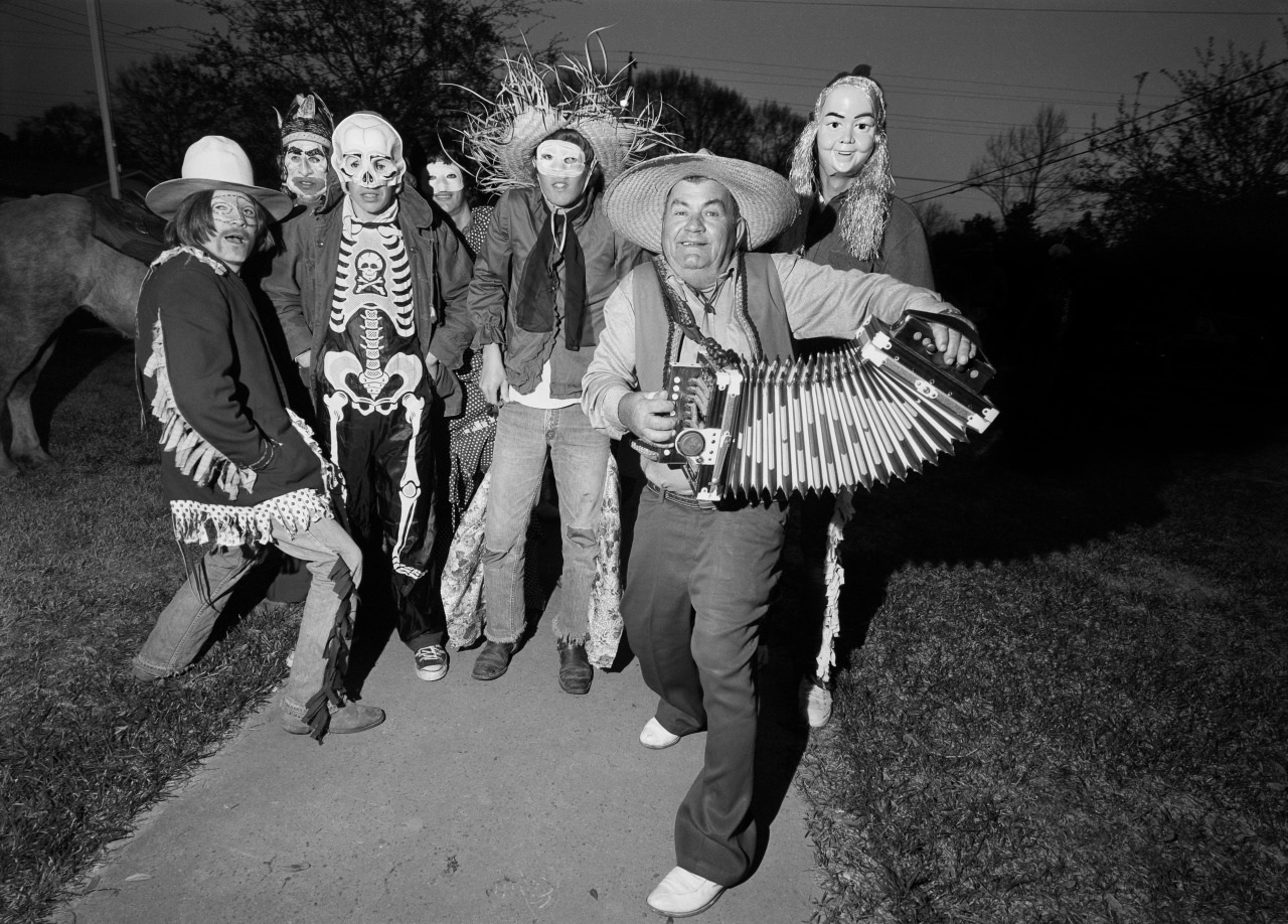 A group of people wearing masks and costumes pose outdoors at night. One person in the foreground plays an accordion. The costumes include a skeleton, straw hats, and various masks. Trees and power lines are visible in the background.