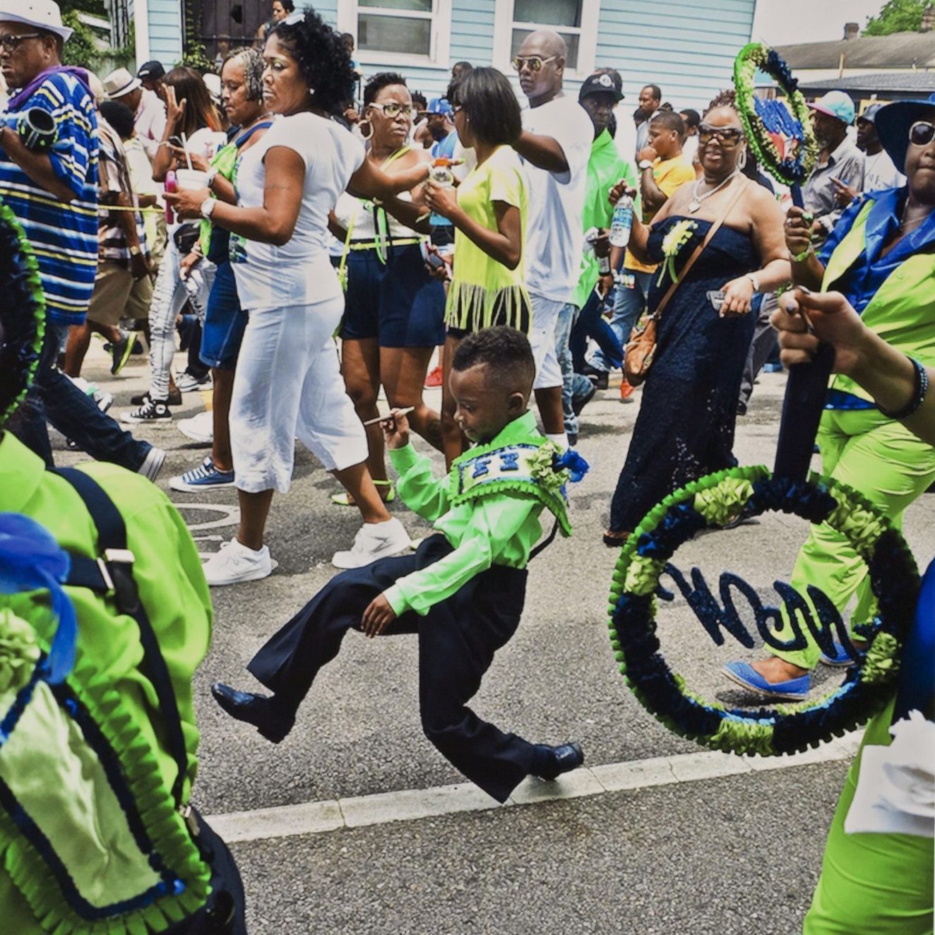A child in a vibrant green outfit energetically dances in a street parade, surrounded by adults wearing white and bright colors. The crowd is lively, with some holding decorated hoops. A festive atmosphere fills the scene.