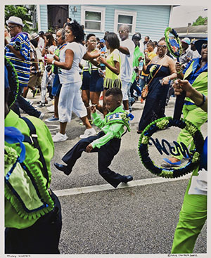 A young boy in a bright green and blue outfit energetically dances in a street parade, surrounded by a lively crowd dressed in colorful attire. The atmosphere is festive, with onlookers enjoying the performance.