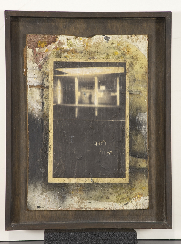 A framed artwork featuring a weathered and abstract background with a central rectangular focal point. The central image contains blurred shapes and the handwritten words I am him. The outer border is dark and rustic.
