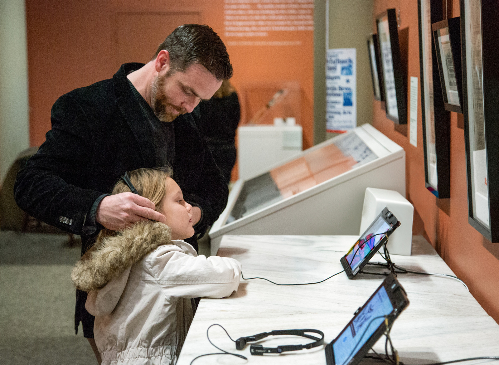 A man helps a young girl adjust her headphones at an interactive exhibit in a museum. They stand by a display with screens and audio equipment, focused on the experience. The background features informational plaques on the wall.