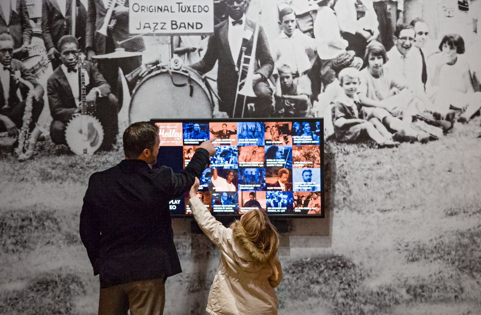 A man and a child interact with a digital display featuring historical jazz images and information. A large black-and-white mural of a jazz band is visible in the background.