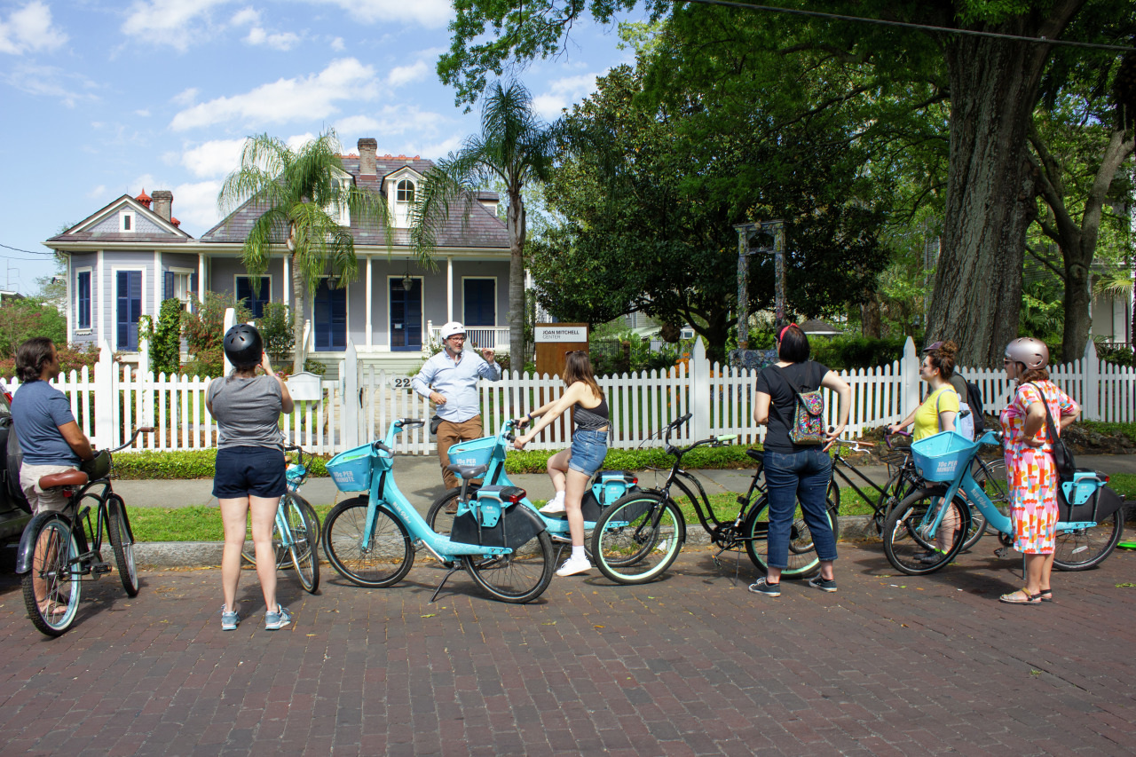A group of people are standing with rented blue bicycles on a brick-paved street. They are listening to a tour guide in front of a historic house with white picket fencing and lush greenery under a partly cloudy sky.