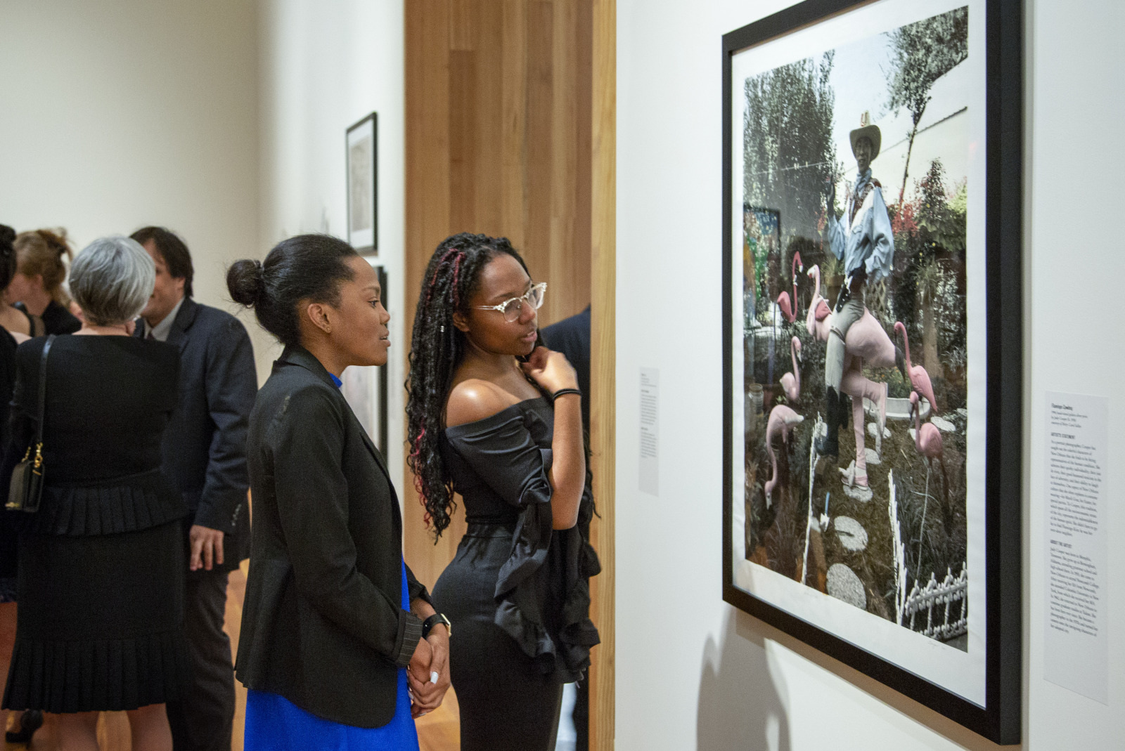 Two women in elegant dresses admire an art piece featuring a person with a cowboy hat surrounded by pink flamingos. Other gallery visitors are also viewing the artwork. The setting is a modern, well-lit art gallery.