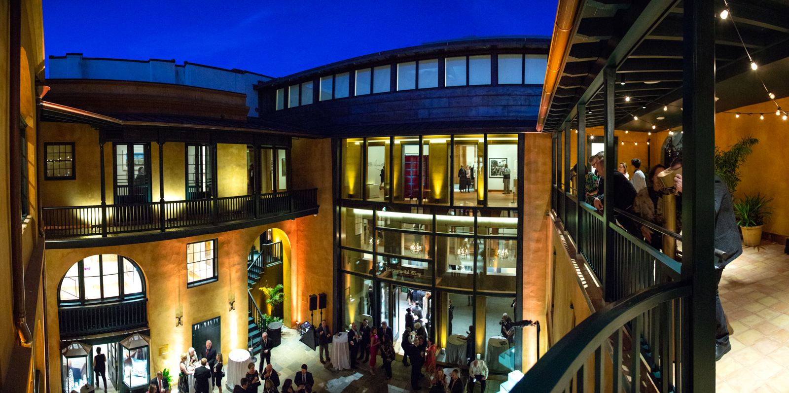 A vibrant courtyard at night, with warm lighting illuminating a two-story building. People are mingling on both levels, enjoying the open-air atmosphere. String lights add a festive touch to the elegant architecture.