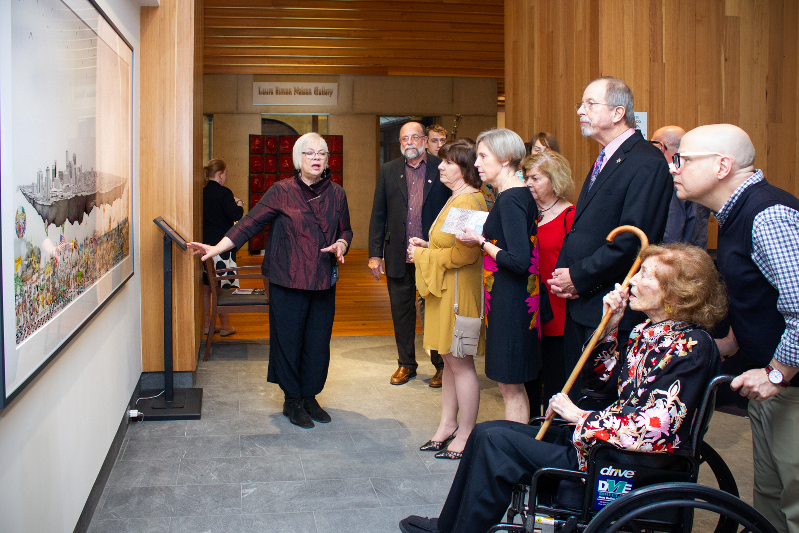 A group of people in a gallery observes a woman explaining a large artwork displayed on the wall. The room has wooden walls, and a woman in a wheelchair holds a cane. Various people wear suits, dresses, and casual attire.