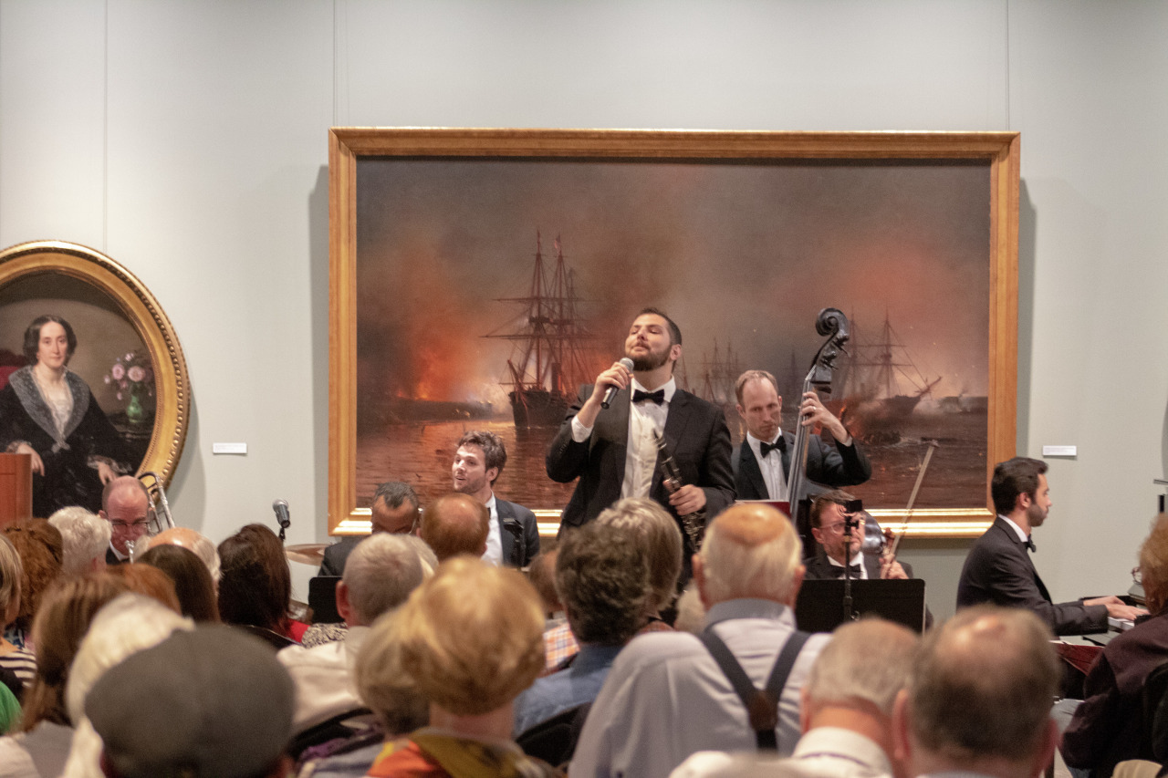 A band performing in a gallery with a large, dramatic maritime painting in the background. The lead singer holds a microphone while other musicians play instruments. An audience watches, seated closely together.