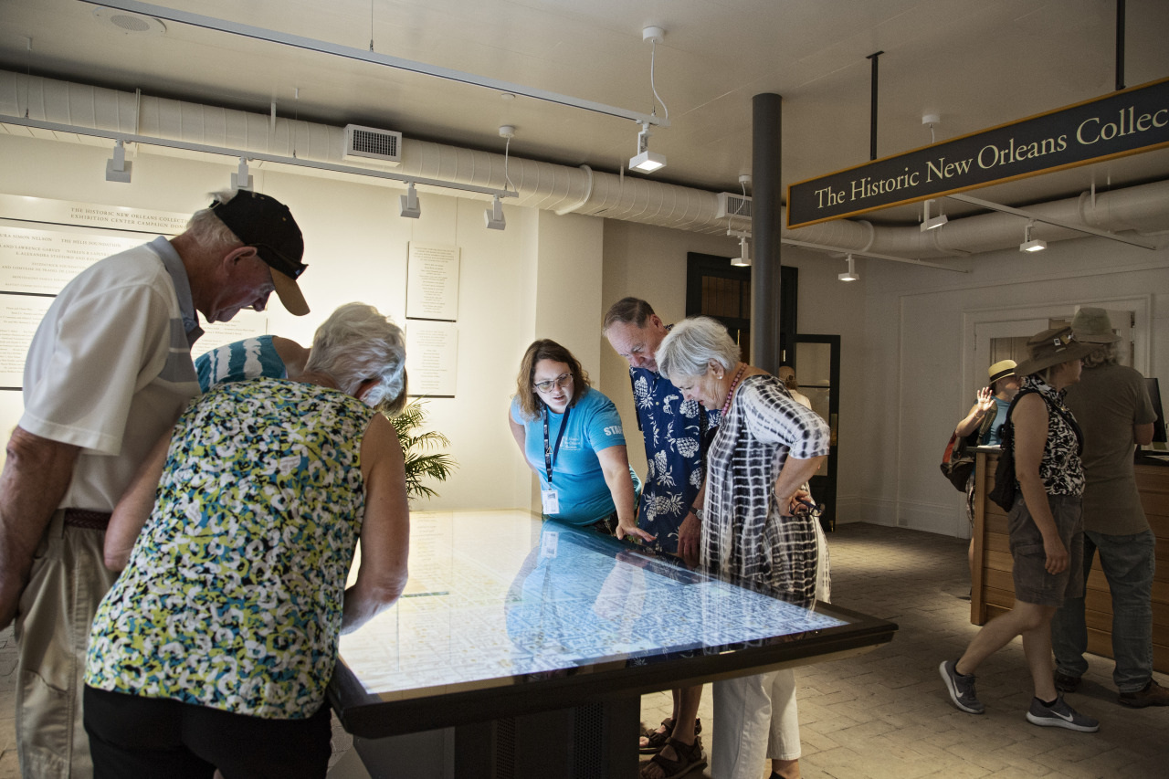 A group of people gather around a large touchscreen table, examining its display. The setting is a museum or historic exhibit with The Historic New Orleans Collection sign in the background. Some visitors wear casual attire and hats.