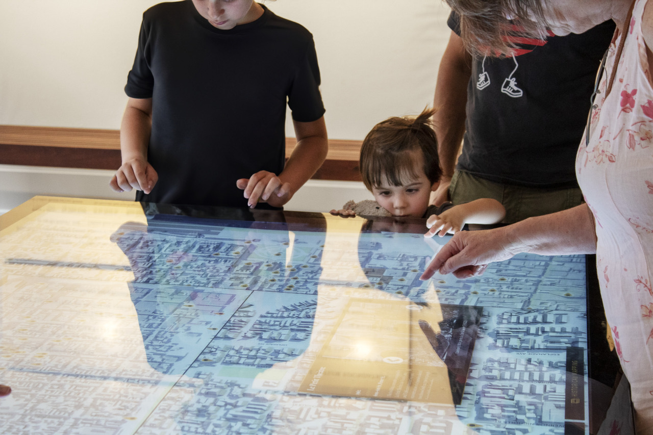 A group of people, including a young child and two adults, are gathered around an interactive touchscreen table displaying a digital city map. The adults are touching the screen, while the child looks on with curiosity.