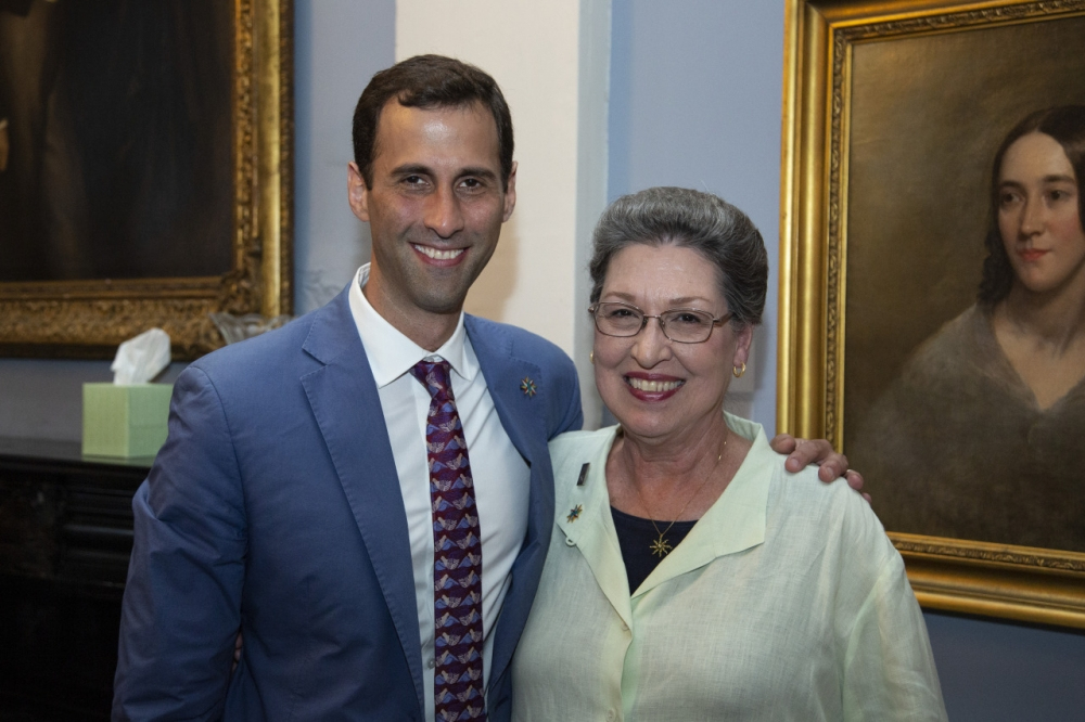 A man in a blue suit with a patterned tie stands smiling next to an older woman in a light blouse. They are both in front of framed paintings, and the setting appears to be an art gallery or museum room.