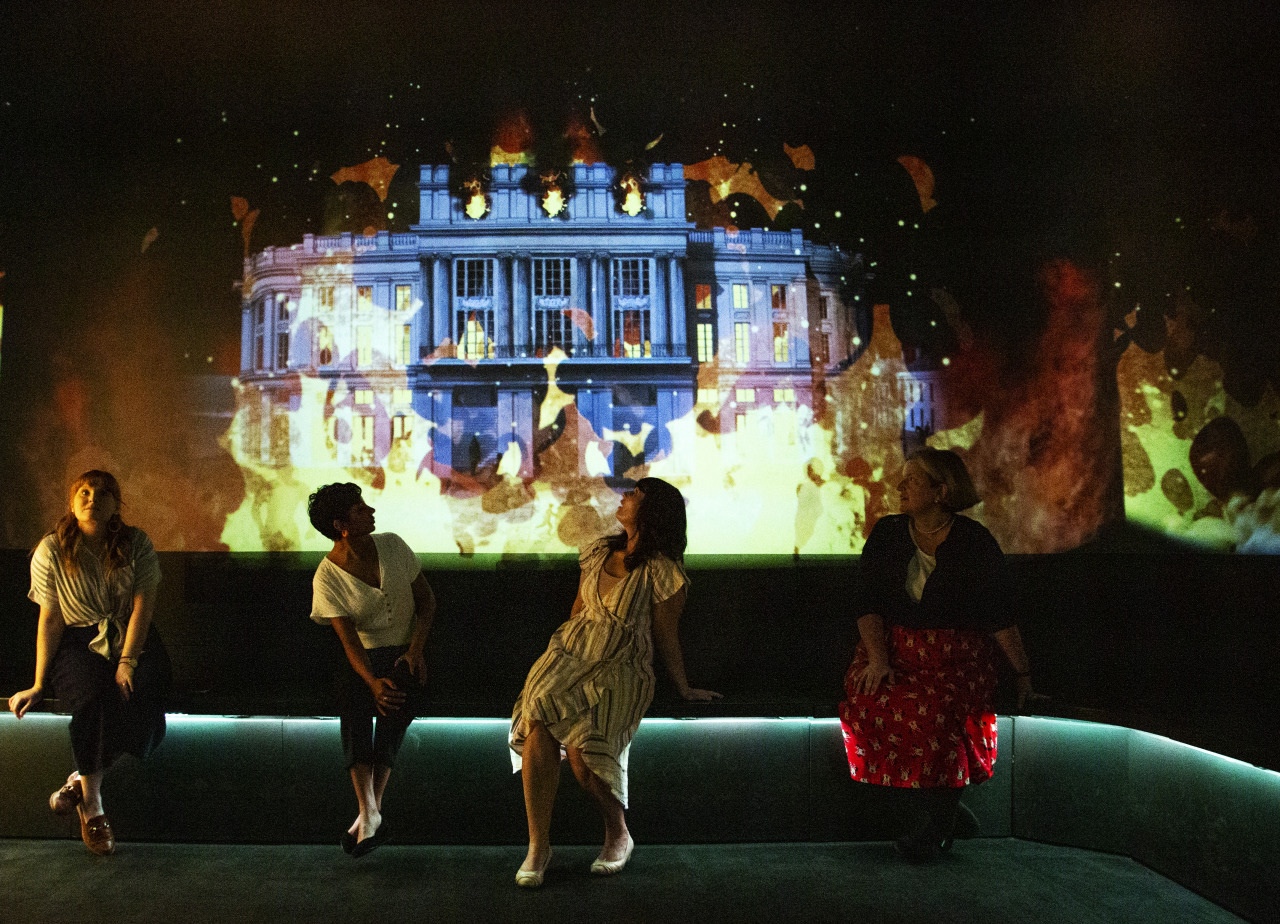 Four people sit on a low wall in a dark room with a vibrant art projection in the background. The projection features a large building surrounded by colorful, abstract designs. The people are casually dressed and appear engaged with the display.