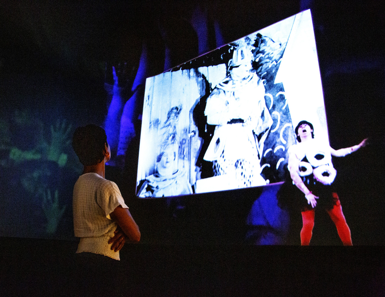 A person stands in a dark room, looking at a large screen displaying an artwork. The screen shows a figure in colorful clothing next to a monochrome abstract image. The background features blue and green shapes.