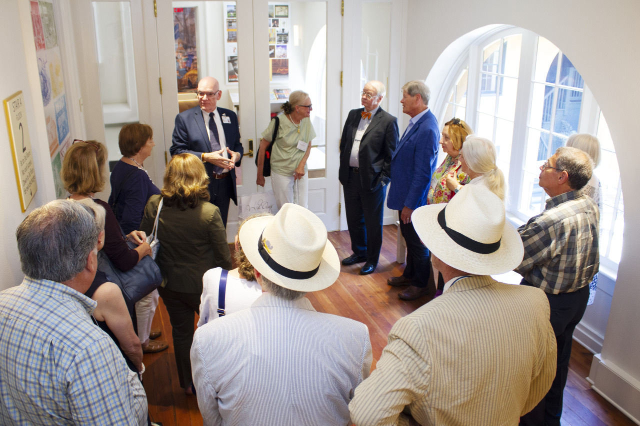A group of people gathered in a room, some wearing hats, listening to a speaker holding a book. They stand on wooden flooring with bright natural light coming through large arched windows. Posters adorn the walls. The mood appears attentive and engaging.
