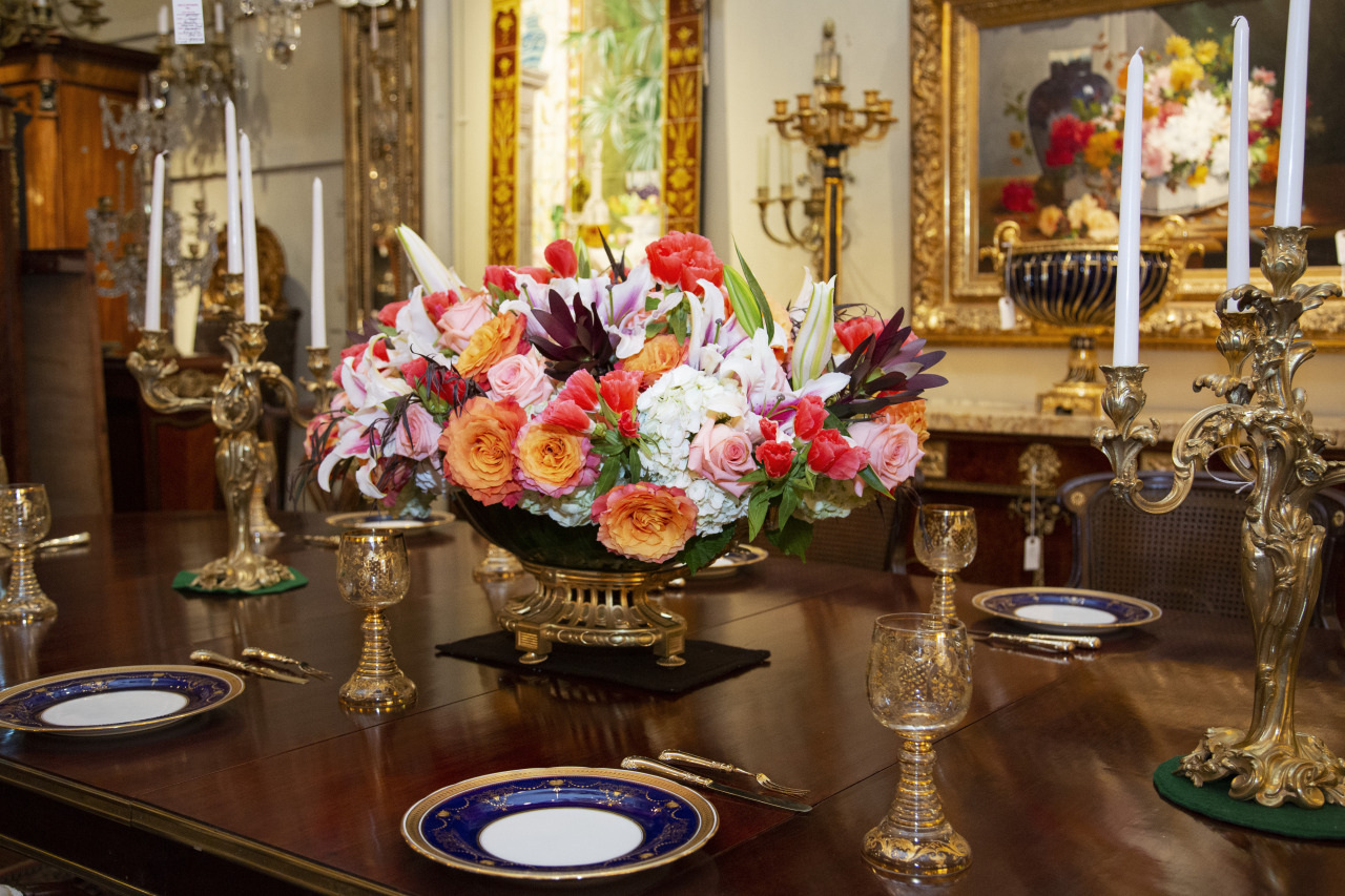An elegant dining room table set with ornate gold candlesticks, crystal glassware, and fine china plates on a dark wood surface. The centerpiece is a lavish floral arrangement with pink, orange, and white flowers in a decorative vase.