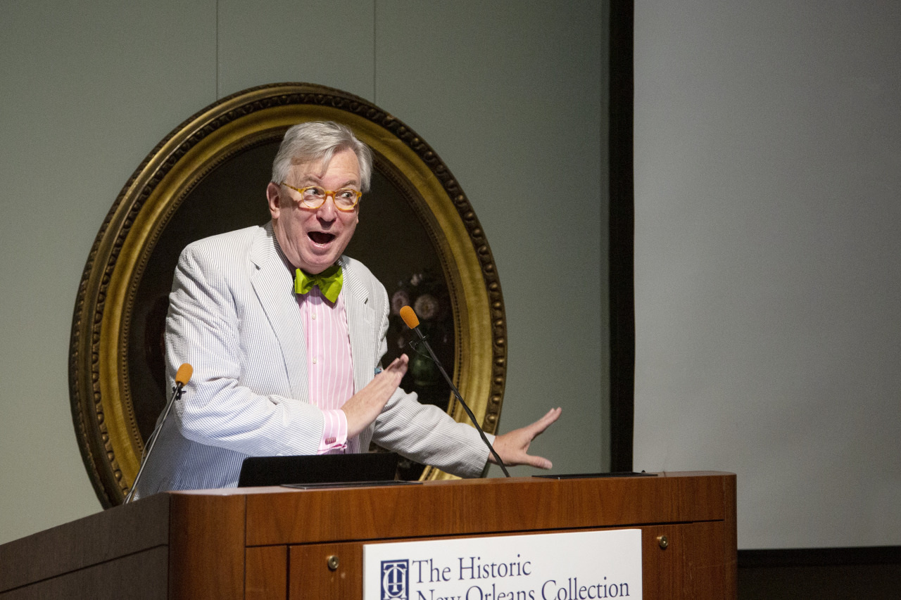 An elderly man with glasses, a green bow tie, and a light suit is speaking energetically at a podium. He gestures with one hand, while the other rests on the podium. Behind him is a large framed portrait.