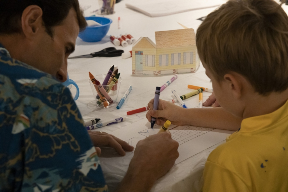 A man and a child are sitting at a table, coloring on sheets of paper with crayons. There are colorful crayons scattered around, and a small model house is on the table. The atmosphere seems creative and focused.