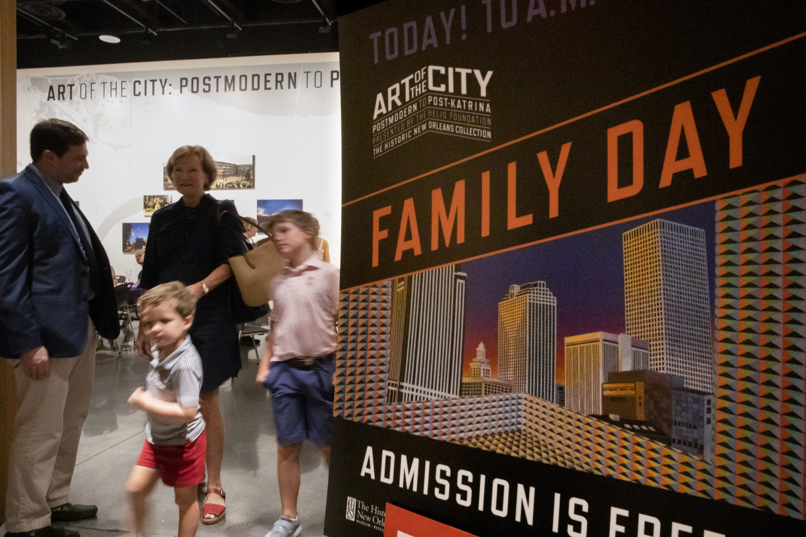 A family stands near a sign promoting Family Day at an art exhibit labeled Art of the City: Postmodern to Present. The sign features city buildings, and the event offers free admission. The family includes two children and two adults.