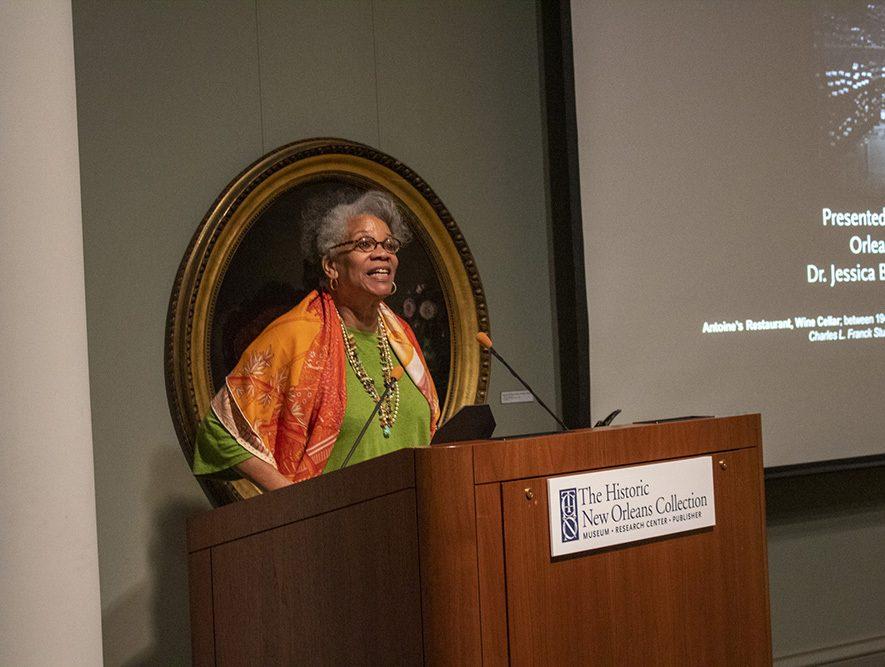 Jessica Harris wearing a colorful shawl stands at a podium, speaking. The podium bears the sign The Historic New Orleans Collection. A projector screen is visible in the background with partially readable text.