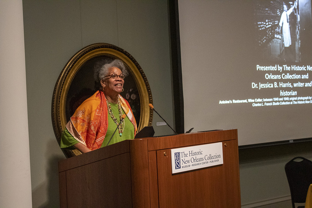 A woman in a colorful shawl speaks at a podium during a presentation. Behind her is a screen displaying text related to the Historic New Orleans Collection and Dr. Jessica B. Harris. There is also a decorative frame on the wall.