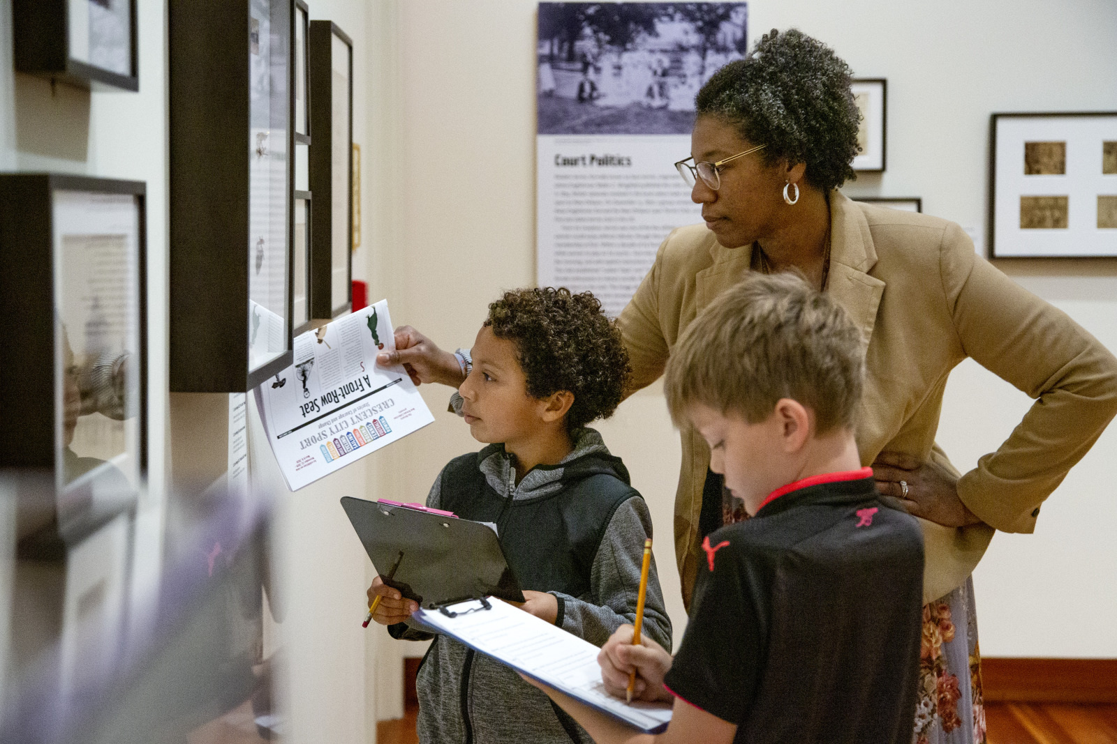 A teacher guides two students who are writing on clipboards in a museum. They are examining an exhibit with photographs and text on the wall. The group is focused and engaged in the educational activity.