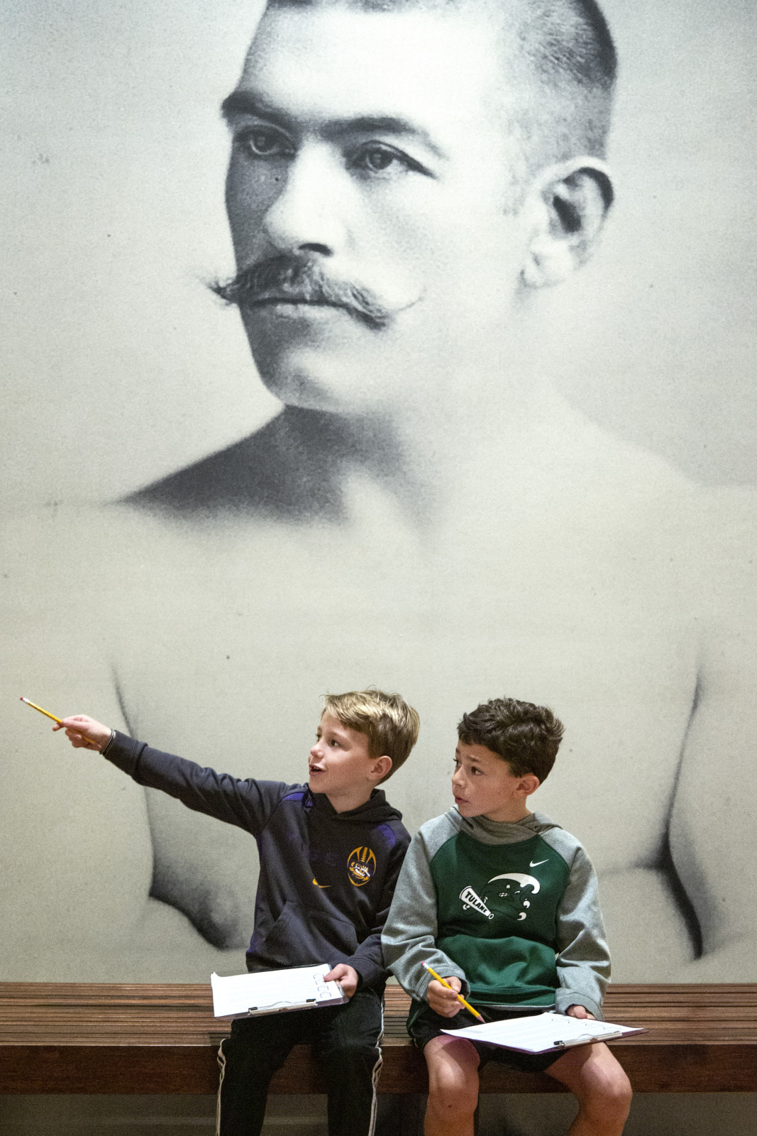 Two young boys sit on a bench with notebooks, one pointing to something off-frame. They are in front of a large black-and-white historical photograph of a shirtless man with a mustache. The boys appear to be engaged in a museum activity.
