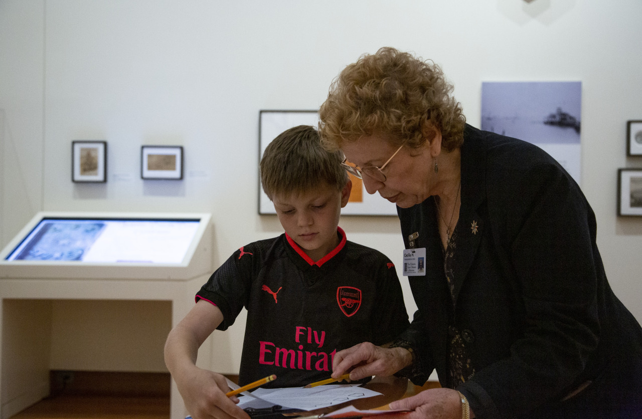 An elderly woman helps a young boy with his work in an art gallery. The boy wears a black and red sports jersey, holding a pencil and paper. Art pieces and a digital display are visible in the background.