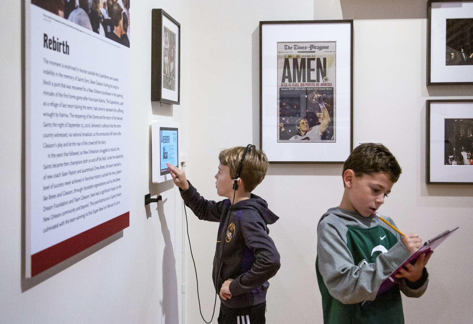Two children in a museum exhibit. One child points at a tablet on the wall while wearing headphones. The other child writes on a notepad. They are surrounded by framed text and images.