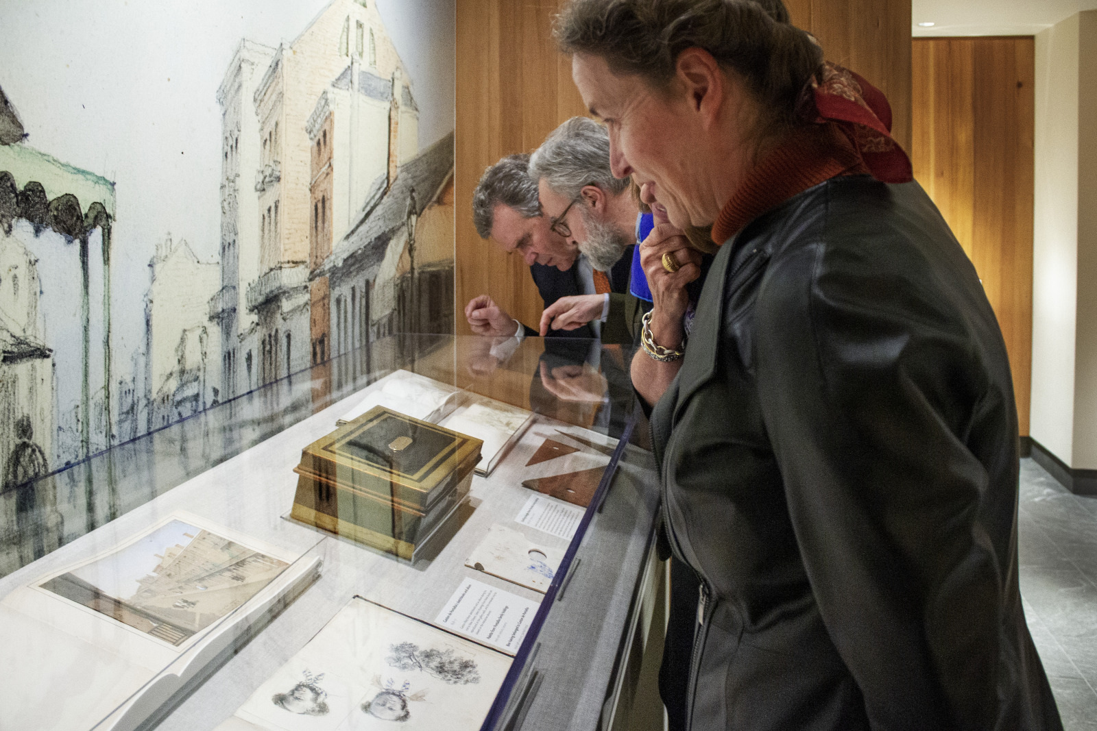 Three people examine historical artifacts and documents displayed in a glass case at a museum. Behind them is a painting of a cityscape, and one person wears a leather jacket with a red scarf.