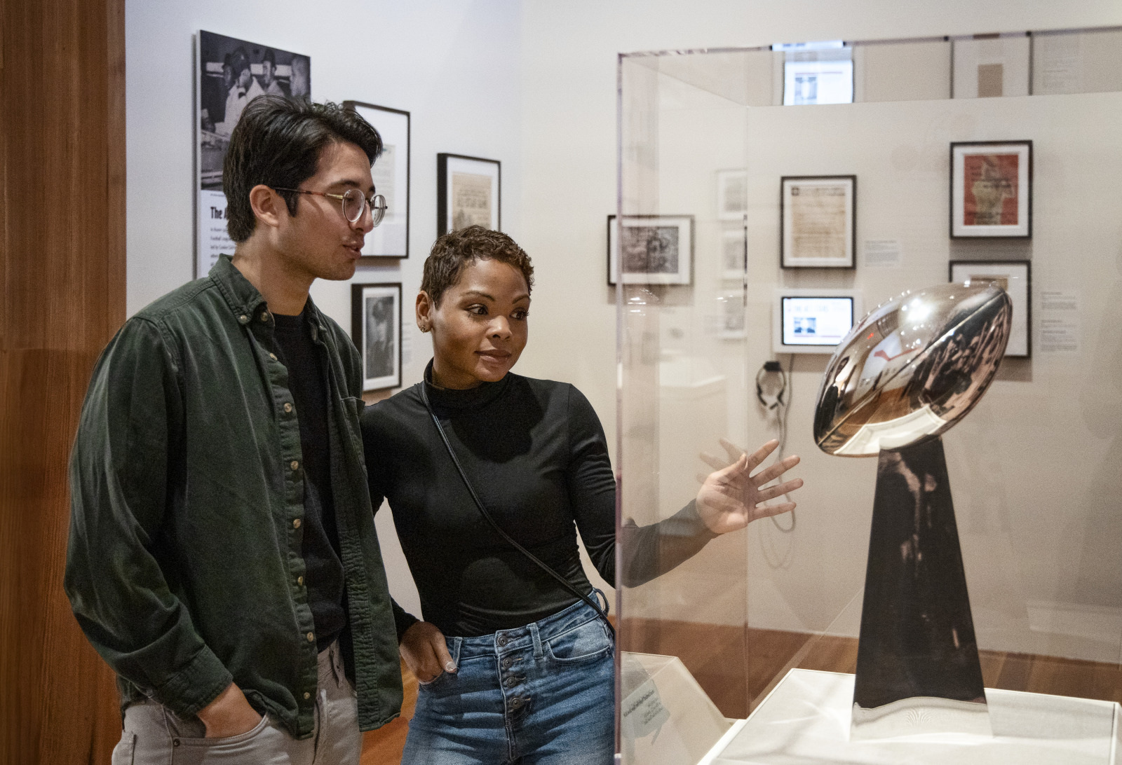 A man and woman stand in an exhibit, looking at a shiny silver trophy displayed in a glass case. The room is decorated with framed photos and documents on the walls. The atmosphere appears quiet and contemplative.