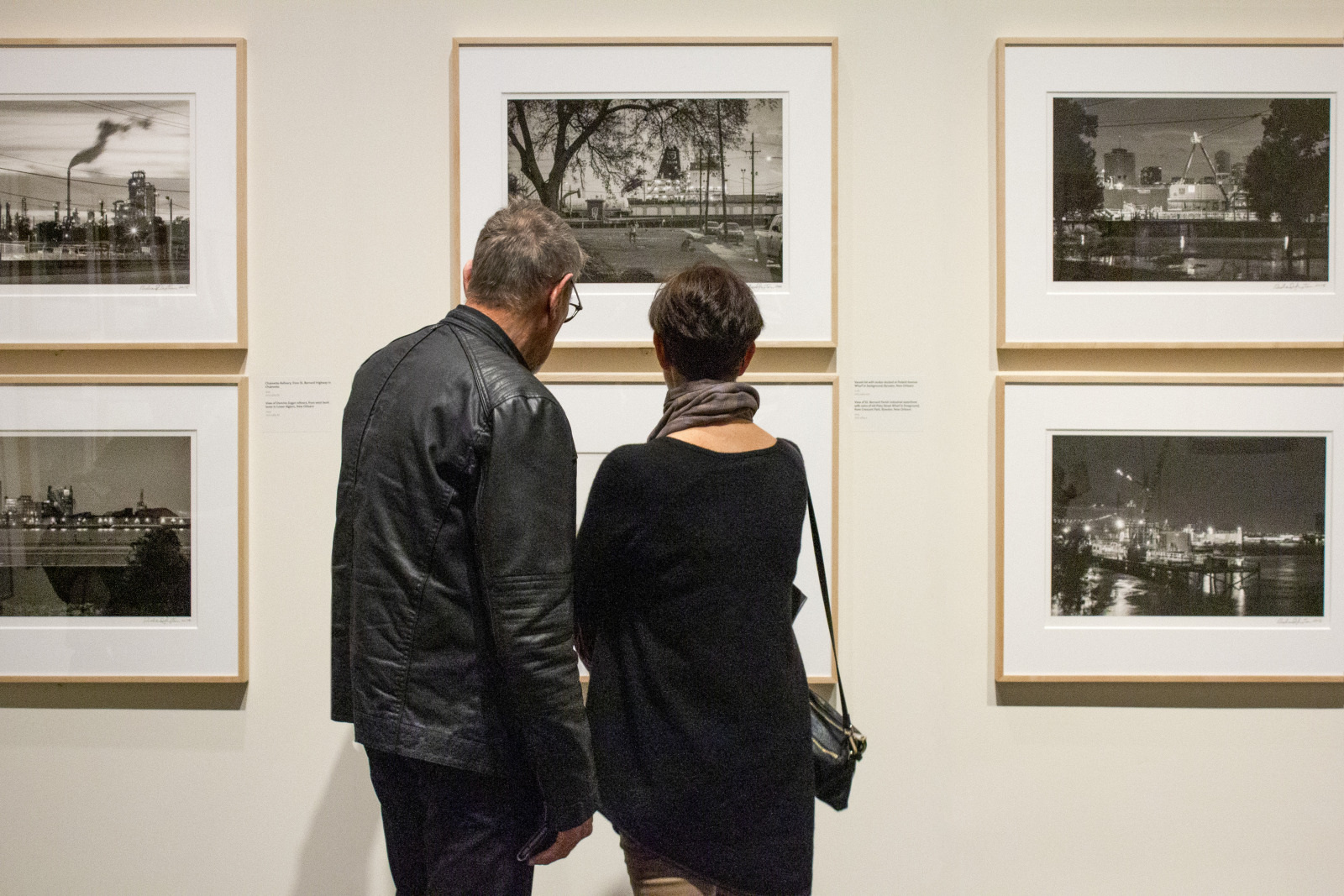 Two people stand close together in front of a gallery wall, looking at framed black and white photographs. The photographs appear to depict various cityscapes and industrial scenes. Both individuals are seen from behind.