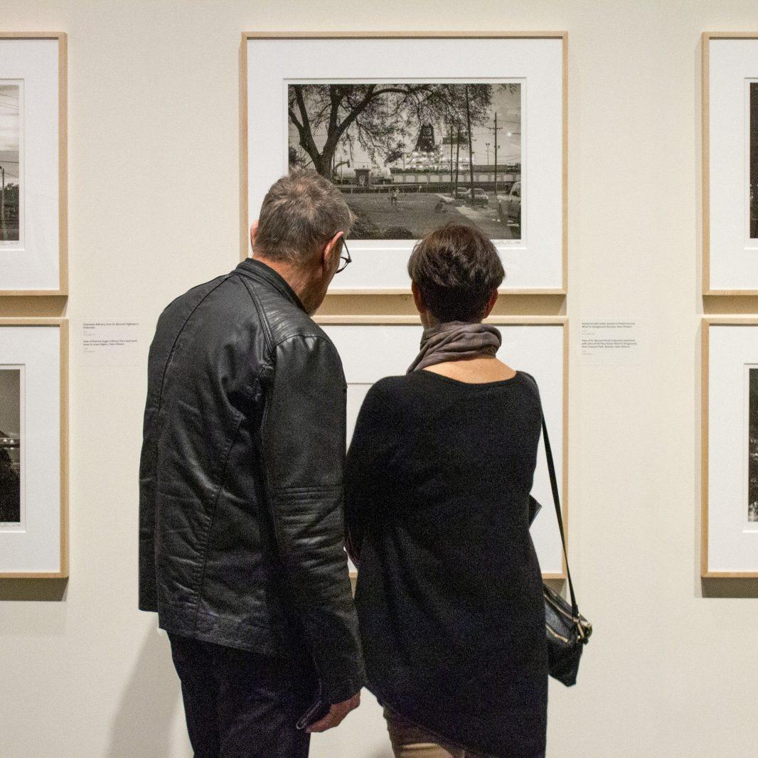 Two people stand closely together, facing framed black-and-white photographs on a gallery wall. Both wear dark clothing; one has a scarf and the other a leather jacket. The atmosphere is quiet and contemplative.
