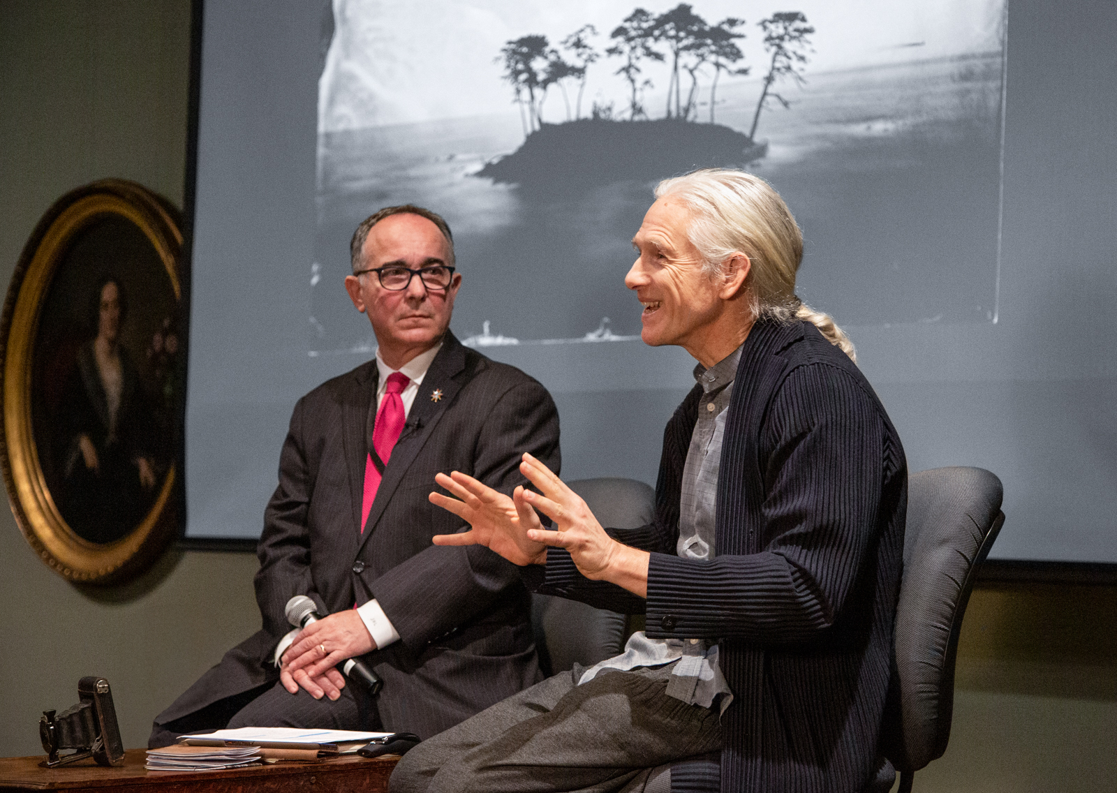 Two men are seated on stage during a discussion. The man on the right gestures while speaking, wearing a casual navy outfit. The man on the left listens attentively, dressed in a suit and red tie. A landscape photograph is displayed on a screen behind them.