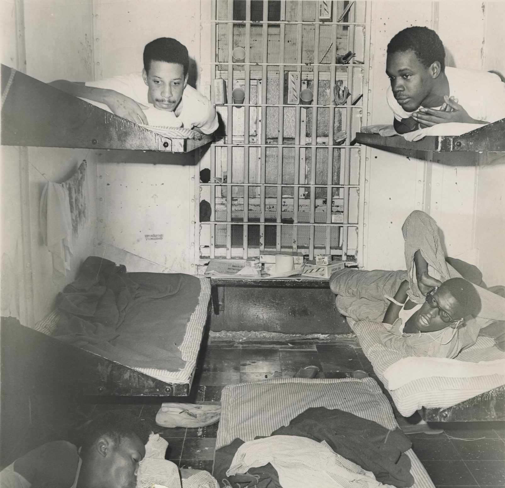 A press photograph of an inmate cell inside Orleans Parish Prison. Three Black men can be seen lying on metal rack beds and another man is partially visible lying on a cot on the ground. Newspapers, empty milk cartons, candy boxes, and cups can be seen on a shelf beneath the window.