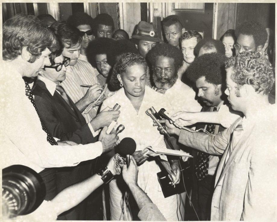 A woman speaks into multiple microphones held by reporters in a crowded room. Several people surround her, some taking notes and others looking on attentively. The image is in black and white.