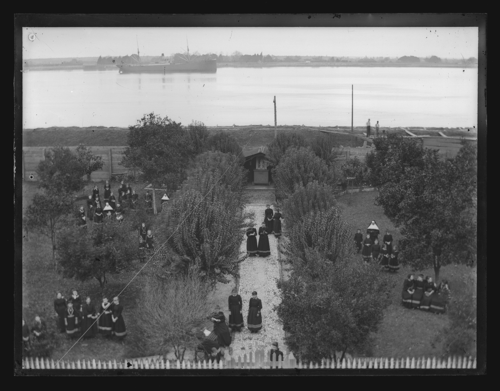 Black and white photo of an elevated view looking down to uniformed pupils standing in groups or walking along a tree-lined path toward a shrine at Ursuline Academy and Convent on Dauphine Street. Several Sisters are shown among the students. The Mississippi River can be seen beyond the tall wooden fence in the distance.