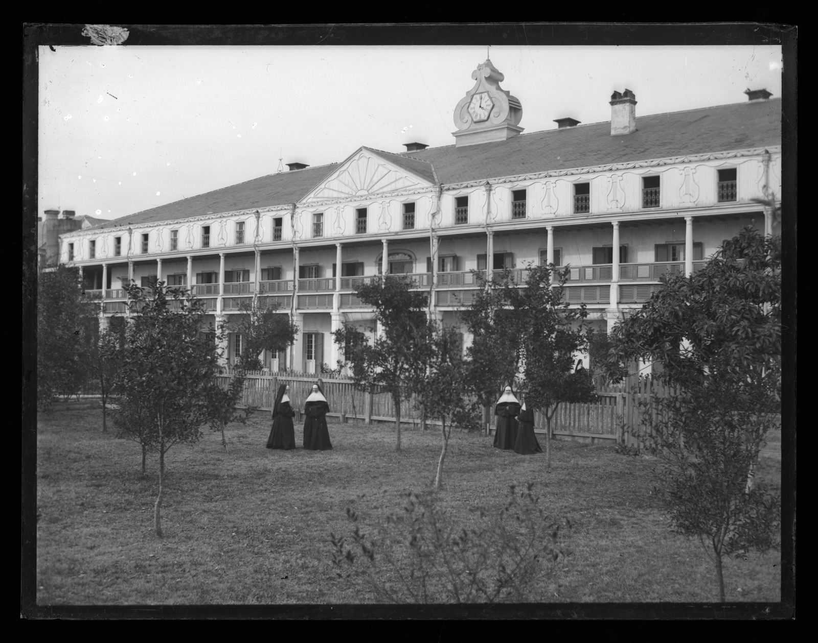 Black and white photo showing a view of four Sisters conversing on the lawn before the main building of the Ursuline Convent on Dauphine Street in the Ninth Ward.