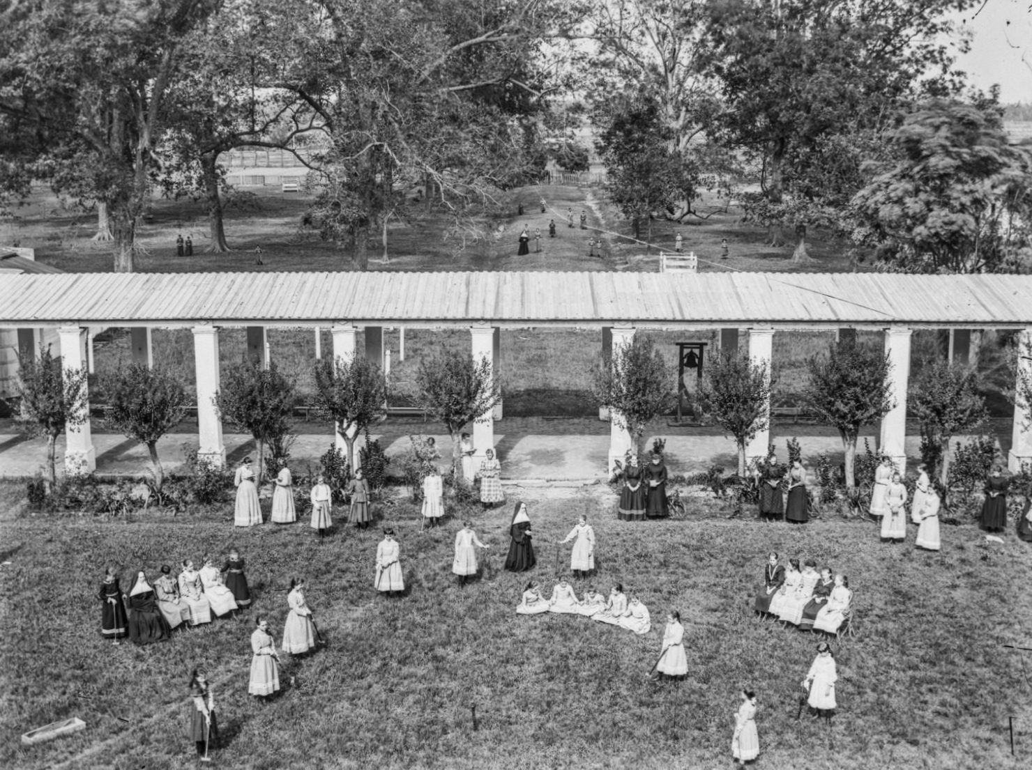 A black and white photo of students and nuns in the academy courtyard at the Dauphine Street Convent in Uptown New Orleans, taken from an elevated position. A walkway crosses the center of the image with a lawn in the foreground and trees in the background.