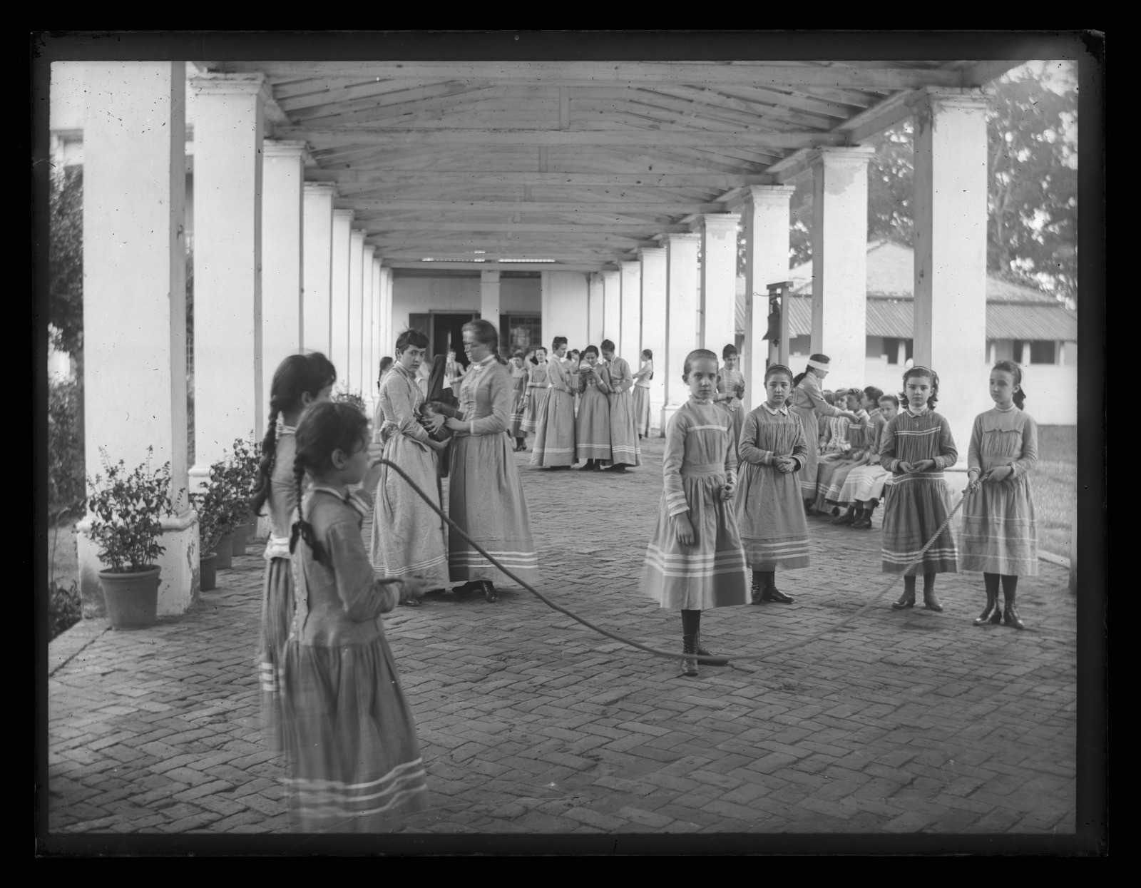 Black and white photo showing a close view of young pupils jumping rope beneath the veranda at the old Ursuline Convent on Dauphine Street. In the background, two young women pass a cat to one another. To the right, a young woman with a blindfold on reaches out toward a student seated on a bench.