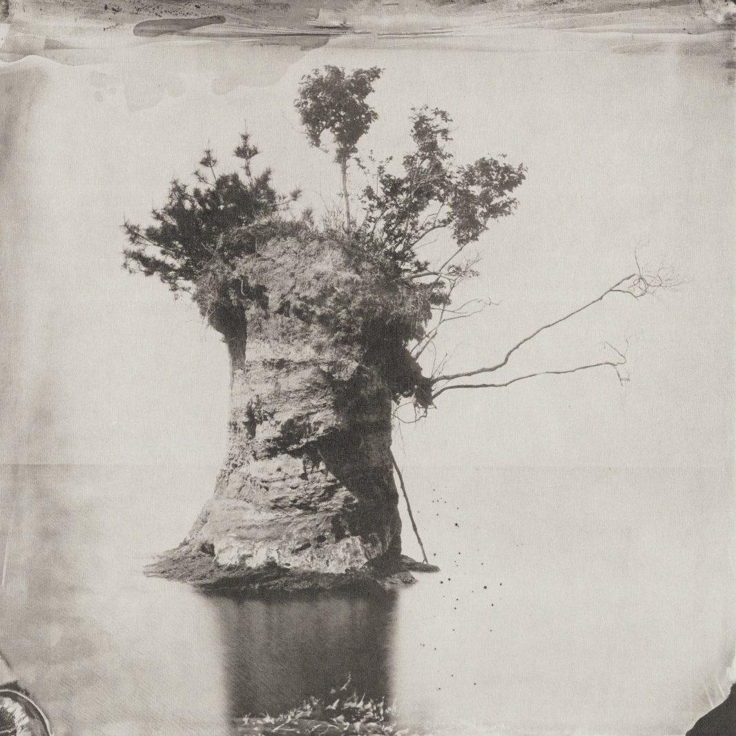 A sepia-toned photograph of a rock island with sparse trees and foliage on top, surrounded by calm water. The island features a solitary, tall tree with branches extending outward. The image has a vintage, textured appearance.
