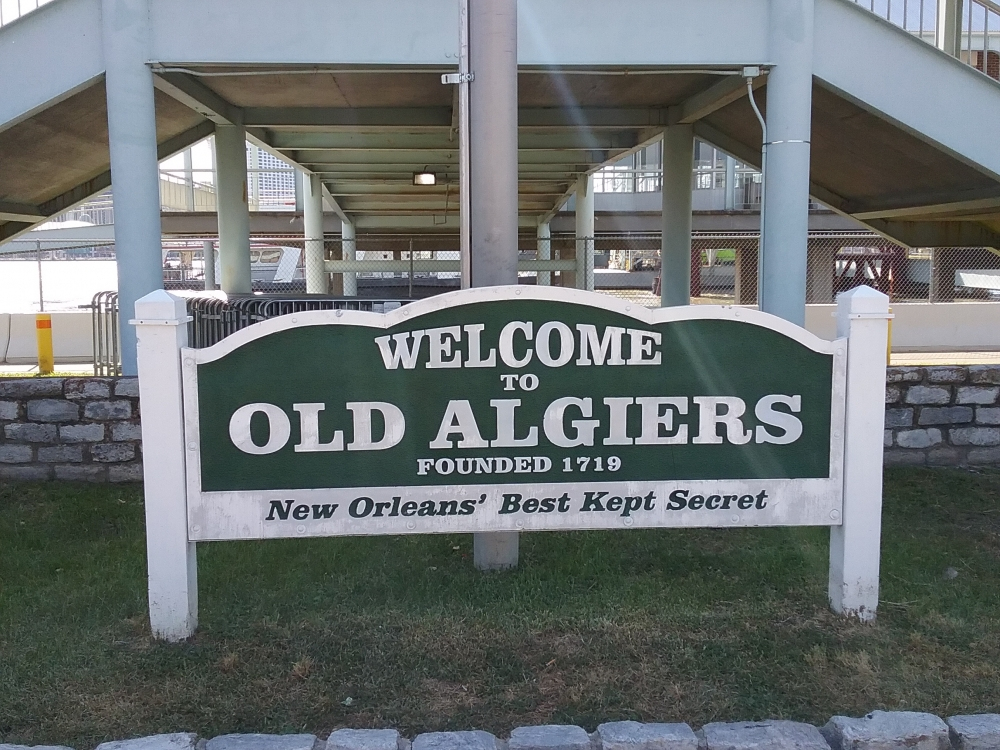 A sign reads Welcome to Old Algiers, Founded 1719. New Orleans Best Kept Secret in front of a building with stairs and a fence, set on a grassy area lined with stones.