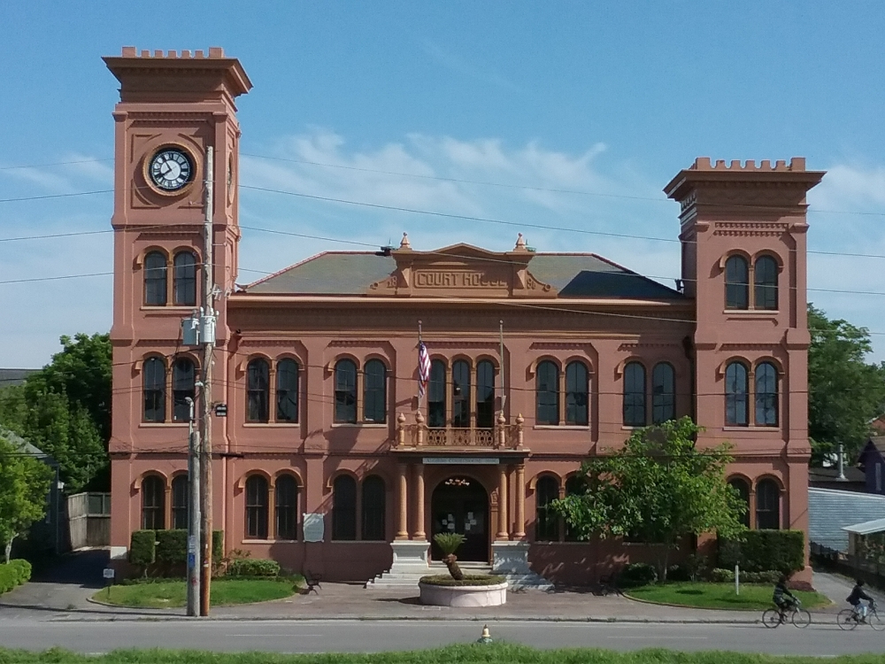 A historic red brick courthouse with two clock towers stands under a clear blue sky. An American flag waves in front, and a cyclist rides past on the street. Lush greenery surrounds the building.