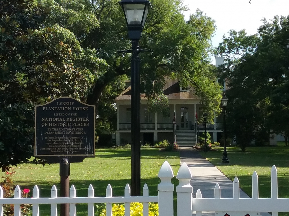 A large historic house is surrounded by trees and greenery. A sign reads Lebeuf Plantation House Listed on the National Register of Historic Places. A white picket fence and a paved path lead to the homes entrance.
