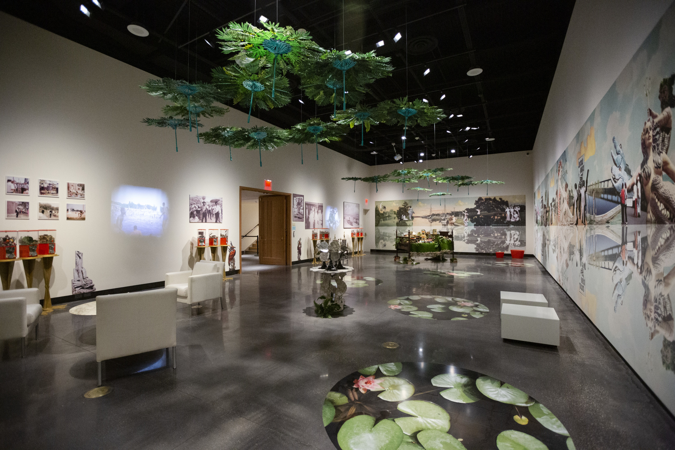 A modern exhibition space with white walls showcases art and photographs. Suspended green installations and lily pad designs decorate the ceiling and floor. White seating is arranged around the room, with a wooden door visible in the background.