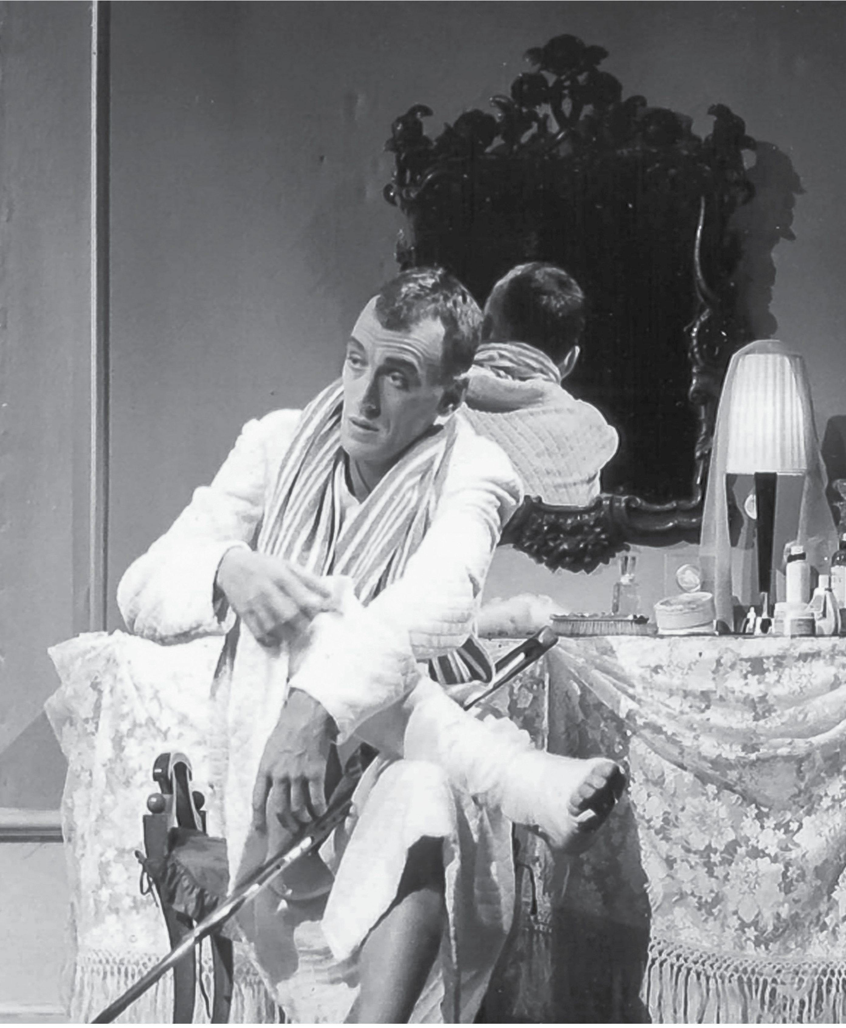 A person in a bathrobe sits on a table, adjusting a bandaged leg. A mirror behind shows their reflection. Nearby is a lamp and various items on a lace-covered table. The setting appears to be a cozy room.