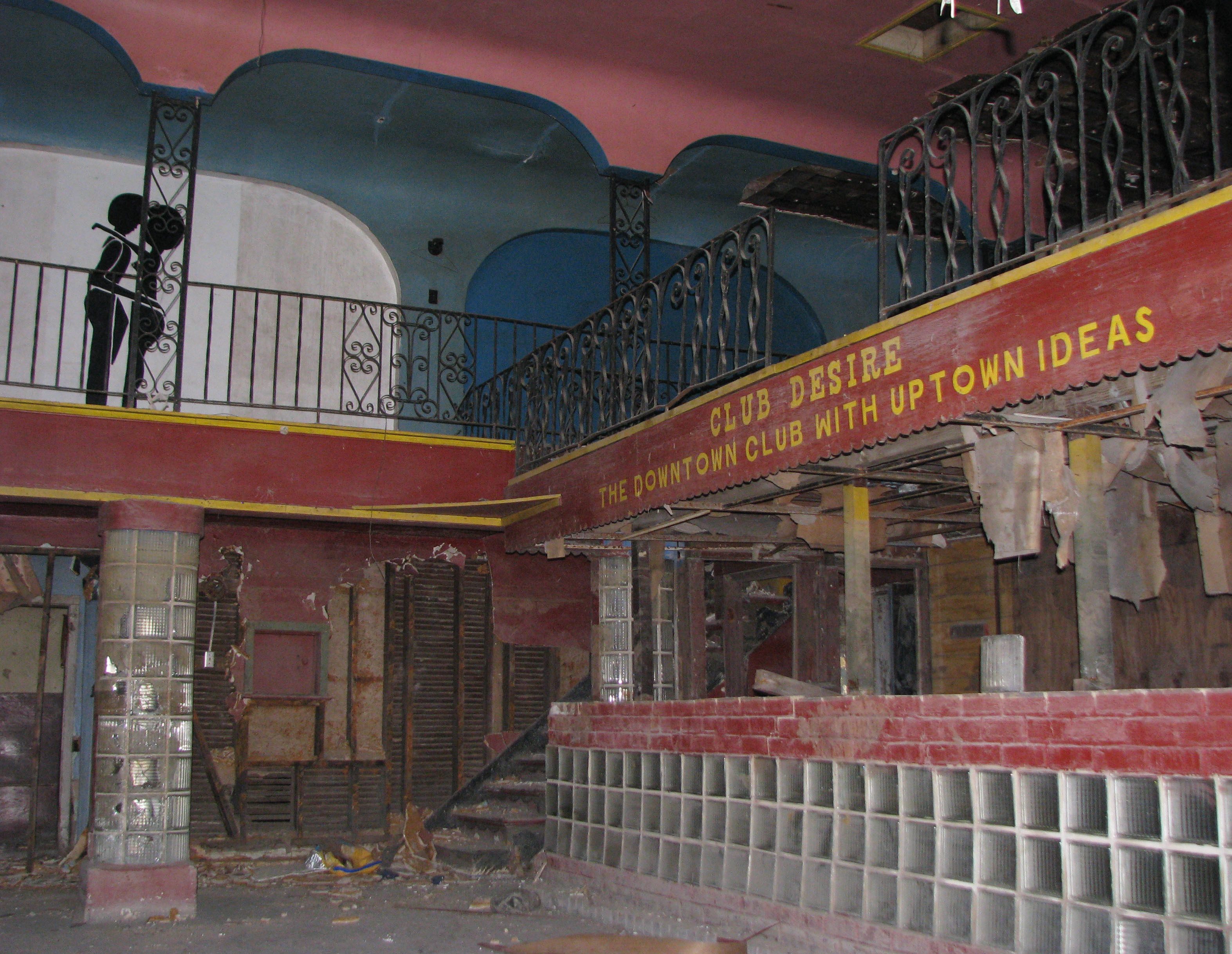The image shows an abandoned nightclub interior with a sign reading Club Desire, The Downtown Club with Uptown Ideas. The space is derelict, with cracked walls, debris, a staircase, and a balcony. A silhouette of a person is on the balcony.
