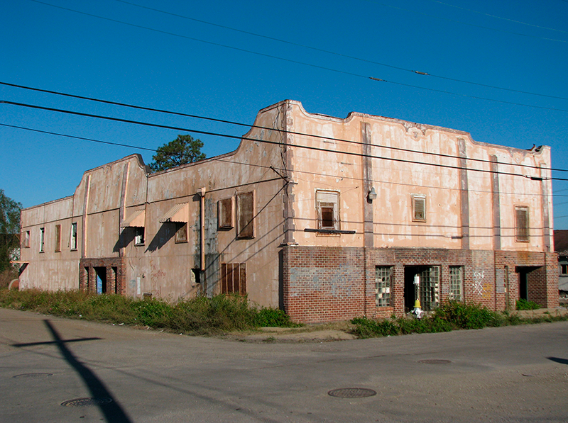 An old, abandoned building with weathered paint and boarded-up windows stands on a street corner. The structure has a mix of brick and stucco walls, and the surrounding area is overgrown with weeds. Power lines are visible in the foreground.